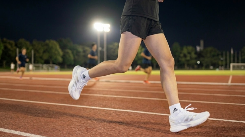 夜の陸上トラックを後ろから捉えた、中学生女子がアディゼロ EVO SLを履いて走る足元フォーカスの写真